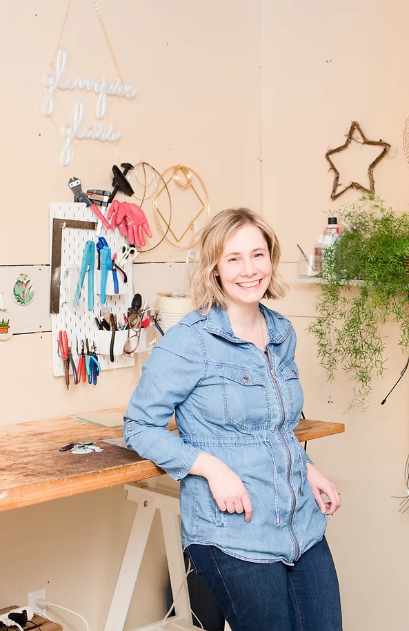 Kara Bussey, founder of Glimpse Glass leans on her workbench in her studio with tools behind her
