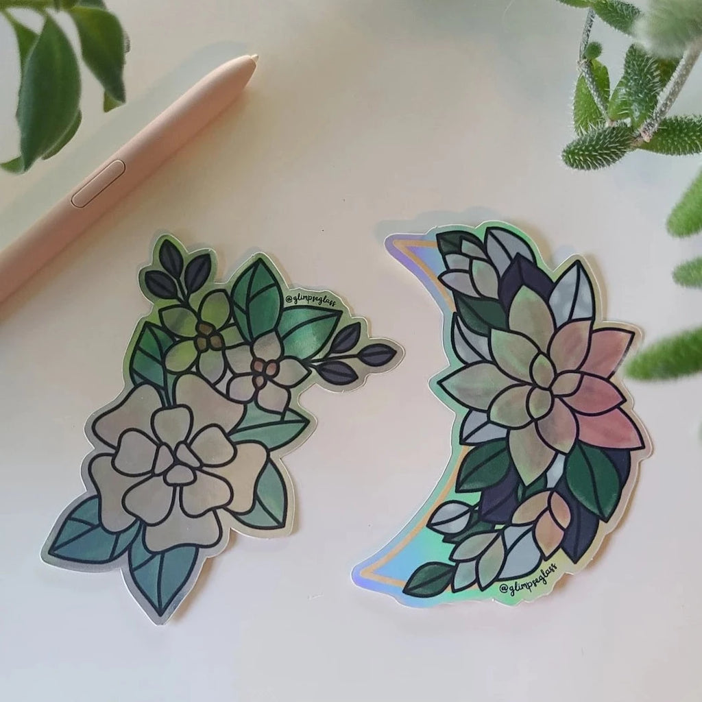 two shiny vinyl stickers, a moon with a pink flower and green leaves, a cream flower with teal leaves on a metallic background